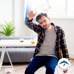 A distressed man sits in a chair, holding an empty alcohol bottle over his head, with a table behind him containing more bottles and a glass of liquor. Why Do People Become Alcoholics? Examining the psychological and emotional reasons behind alcohol addiction.