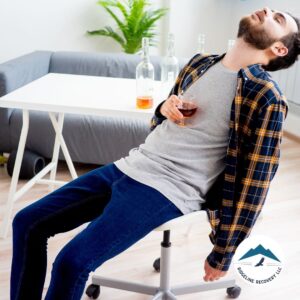 A man in casual clothing slumps in a chair, holding a glass of alcohol, with empty bottles on the table behind him. Why Do People Become Alcoholics? Exploring the emotional, psychological, and social triggers behind addiction.