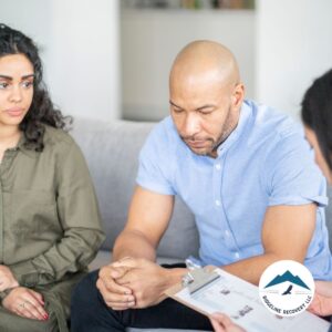 A man sits with his hands clasped, appearing deep in thought, as a counselor reviews paperwork. Sober living house options in Ohio provide essential recovery support, helping individuals transition from rehab to independent living.