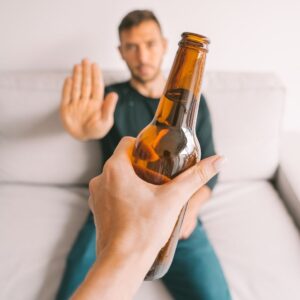 A man sitting on a couch raises his hand to refuse an offered beer, symbolizing the struggle of overcoming Effexor addiction and alcohol dependence.