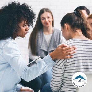 A compassionate therapy session where a woman provides support by placing a comforting hand on a fellow group member’s back. Rehab Facilities That Take Medicaid offer structured group therapy to aid in addiction recovery.