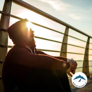 A man in a hoodie sits by a railing during sunset, reflecting on his journey to sobriety, symbolizing the strength and hope found in rehab for couples Ohio to heal together.