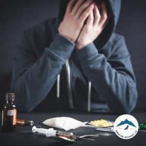 A distressed individual in a hoodie sitting at a table with drugs and paraphernalia, representing the struggles of addiction and the need for rehab for couples Ohio to heal together.