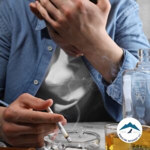 A distressed man sits at a table with a cigarette and a bottle of alcohol, symbolizing the struggles of addiction and the need for Inpatient Programs that provide structured recovery and support.