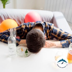 A man in a plaid shirt is slumped over a white table surrounded by alcohol bottles and an ashtray, representing the struggles of addiction and the need for Sober Living Rooms for Rent in Columbus.