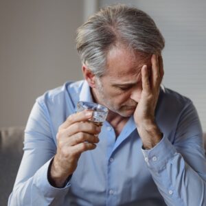 A distressed middle-aged man in a blue shirt sits with a glass of alcohol, holding his head in frustration, illustrating the emotional struggles of Effexor addiction and alcohol dependence.