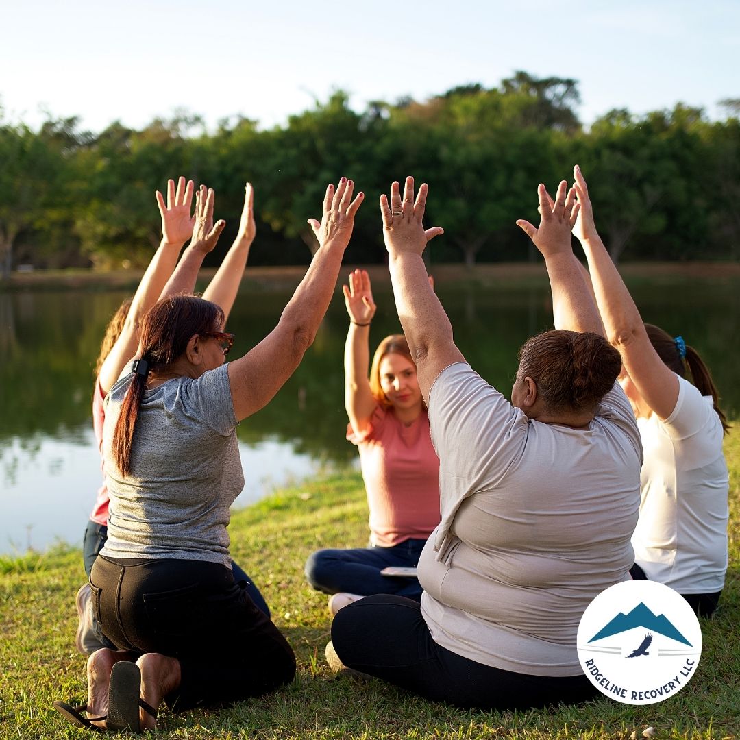 A group of people participates in an outdoor therapy session near a lake, raising their hands together in unity. Free Drug Detox Near Me programs offer holistic healing and community support for addiction recovery.