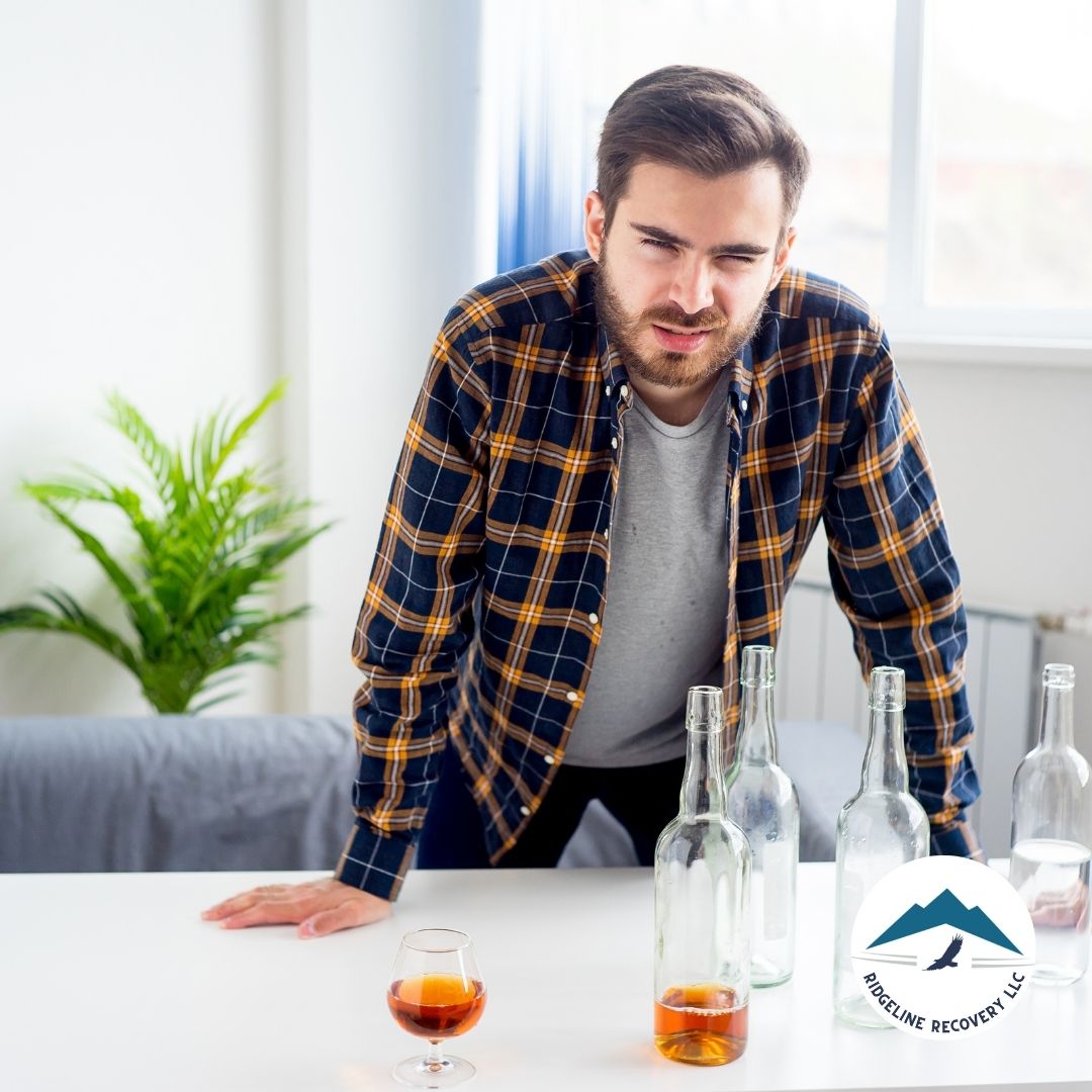 A man in a plaid shirt stands at a table with empty alcohol bottles, looking distressed. This image represents the challenges of sobriety and how recovering alcoholics and dating can be a complex but rewarding journey.