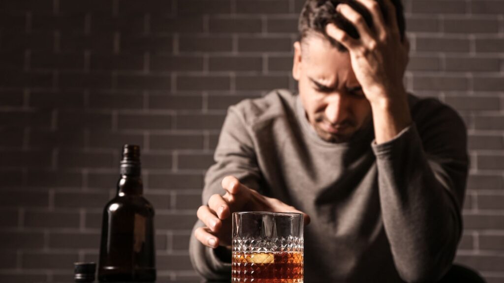 A man overwhelmed with stress and depression sits with a glass of alcohol, reflecting emotional and psychological causes of alcoholism.