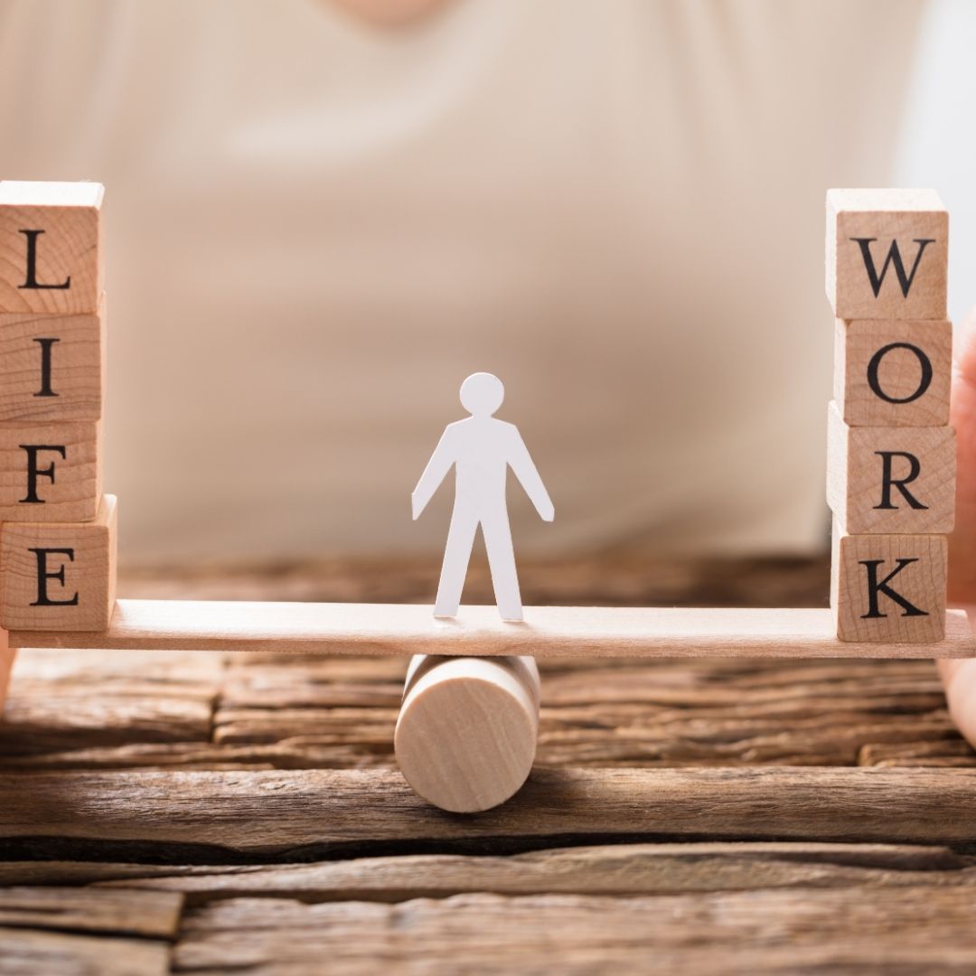 A small wooden balance scale with the words "LIFE" and "WORK" on stacked blocks, with a paper cutout figure standing in the middle, symbolizing Living in Balance for recovery and well-being.
