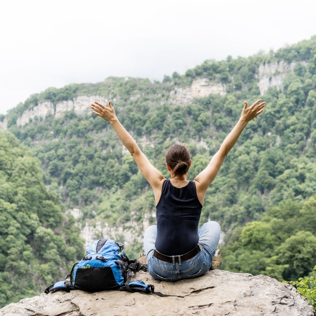 A woman sits on a mountain ledge with her arms raised, embracing nature and fresh air, symbolizing signs that your body is detoxing, such as renewed energy, mental clarity, and overall well-being.