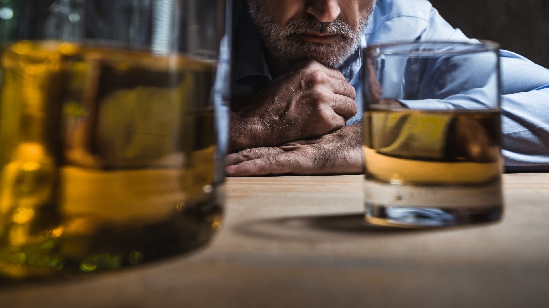 A man sitting alone, drinking alcohol in a dimly lit room, reflecting on his alcohol addiction.