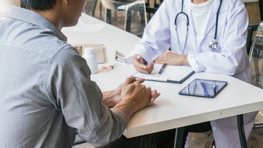 An intimate perspective of a confidential, one-on-one consultation regarding addiction recovery. A patient's hands are clasped, indicating seriousness, while the clinician across the table appears attentive with a clipboard and tablet nearby. The presence of small bottles suggests the medical component of the discussion. This image powerfully represents the combined medical and therapeutic approach of Medication-Assisted Treatment (MAT).